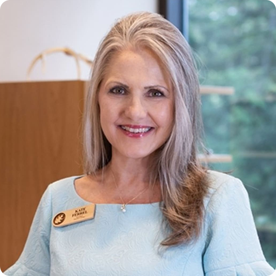 A smiling woman with long, light brown hair, wearing a light blue dress and a name badge, stands indoors in front of a window with greenery outside.