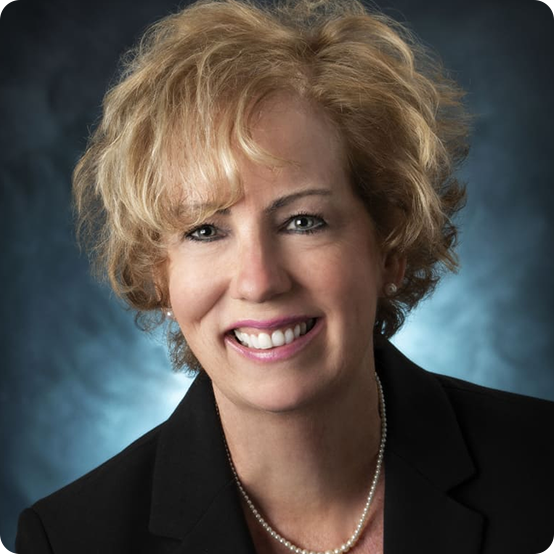 A smiling woman with short, curly blonde hair wearing a black blazer, white pearl necklace, and light makeup, posed in front of a dark, softly lit background.