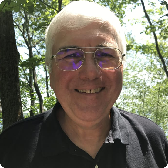 An older man with short white hair, glasses, and a black collared shirt smiles outdoors, surrounded by green trees and sunlight filtering through the leaves.