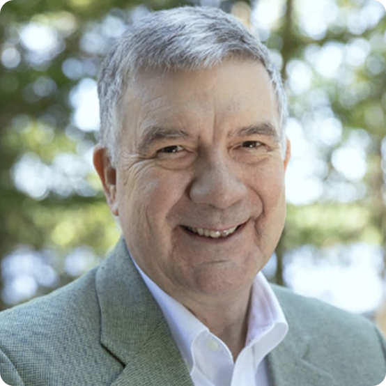 A smiling older man with short gray hair, wearing a light gray blazer and white shirt, stands outdoors with trees and sunlight in the blurred background.
