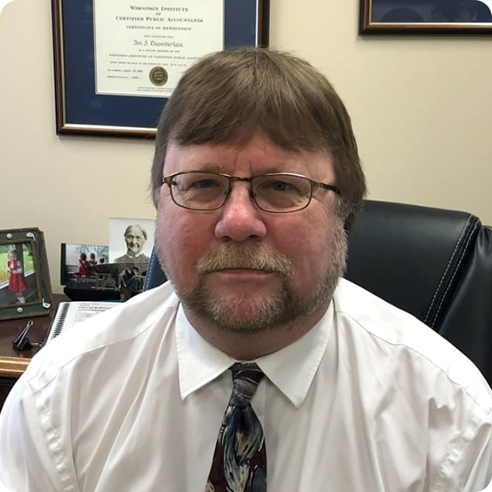 A middle-aged man with glasses, mustache, and beard sits in an office, wearing a white shirt and patterned tie. Behind him are framed certificates on the wall and family photos on his desk.