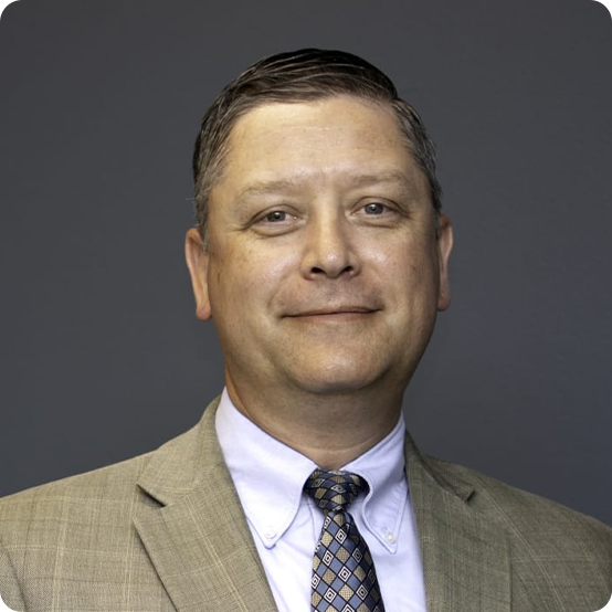 A middle-aged man with short brown hair wearing a beige suit, white dress shirt, and patterned tie, smiling slightly in front of a plain gray background.