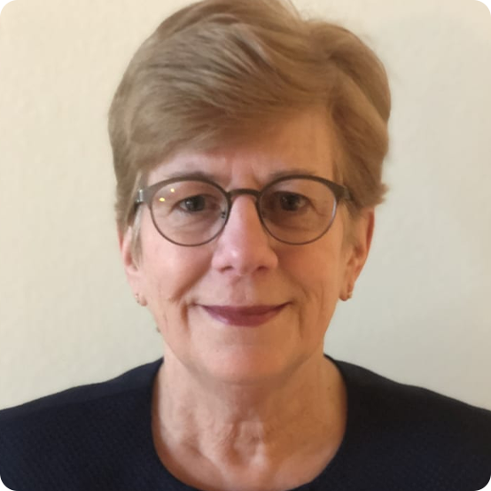 A middle-aged woman with short light brown hair, glasses, and a dark top, smiling slightly in front of a plain light-colored background.