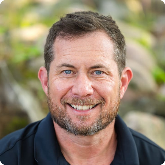 A smiling man with short brown hair and a beard, wearing a dark collared shirt, sits outdoors with a blurred natural background.