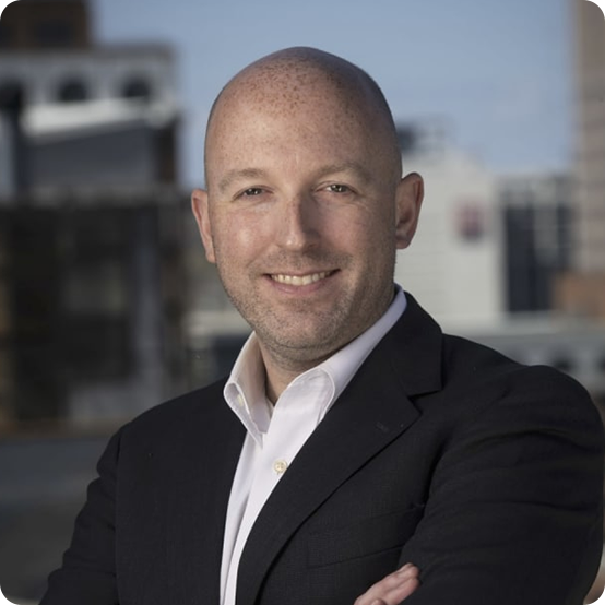 A smiling bald man in a dark suit and white shirt poses outdoors with his arms crossed, with an urban cityscape blurred in the background.