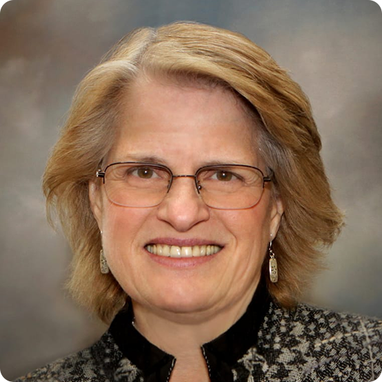 A smiling older woman with short, light brown hair and glasses, wearing a patterned jacket and dangling earrings, poses in front of a blurred grey background.