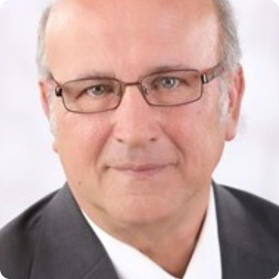 A middle-aged man with light skin, wearing glasses, a dark suit, and a white shirt, posing for a professional headshot against a plain, light-colored background.