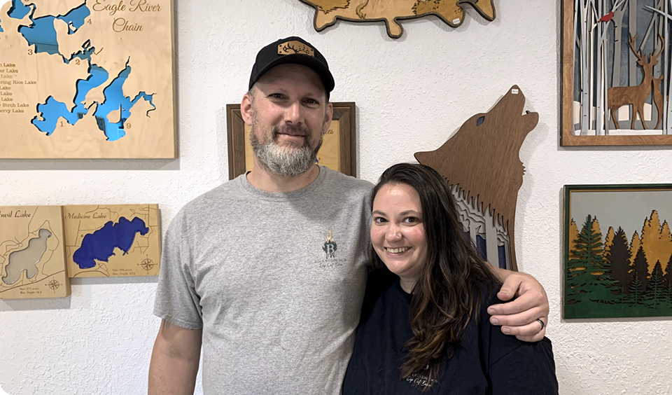 A man and woman stand close together, smiling at the camera. The man wears a gray t-shirt and black hat; the woman has long brown hair and a navy shirt. Behind them are wooden art pieces featuring animals, lakes, and nature scenes.