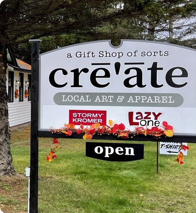 A sign in front of a building reads create: a Gift Shop of sorts, LOCAL ART & APPAREL with Stormy Kromer and Lazy One logos, decorated with autumn leaves. A smaller sign below says open.