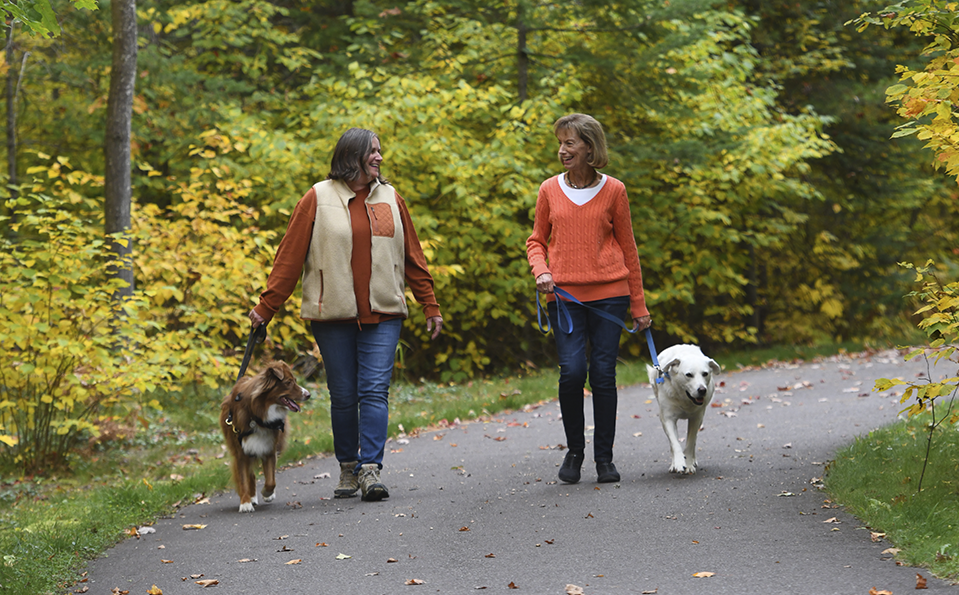 Two women walking dogs on a paved path through a forest with autumn foliage. One woman holds a brown dog’s leash, the other a white dog’s leash. Both women wear sweaters and smile as they walk.