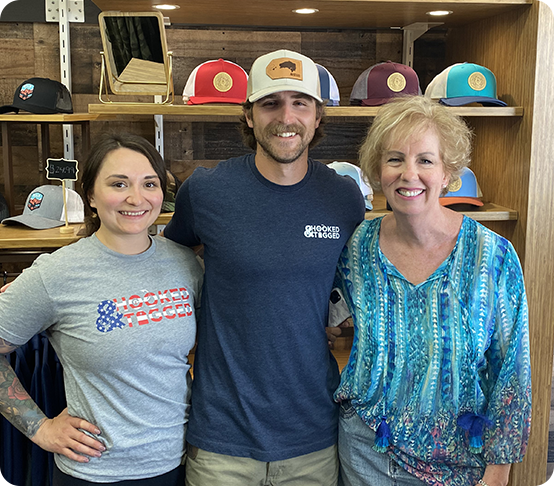 Three people smile while standing together in front of wooden shelves displaying hats. The man in the middle wears a cap and blue T-shirt, and the two women on either side wear casual tops.