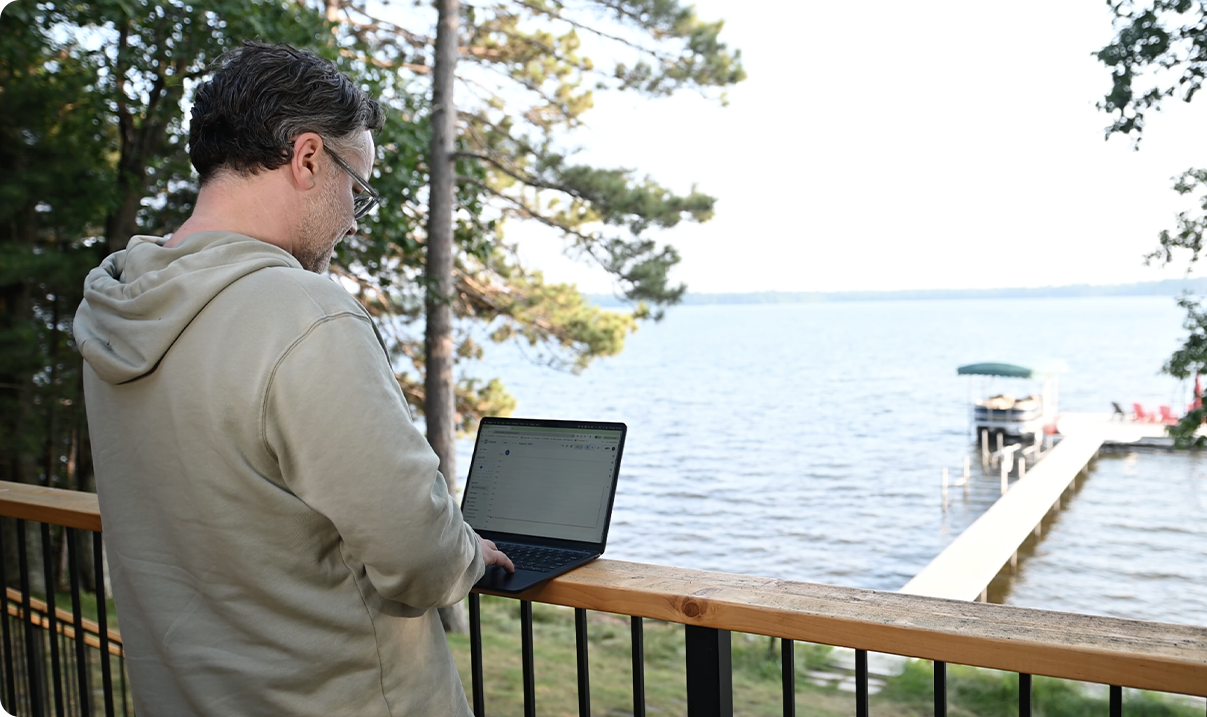 A remote worker in a hoodie types on a laptop while standing on a balcony overlooking a lake, with trees and a dock extending into the water in the background.