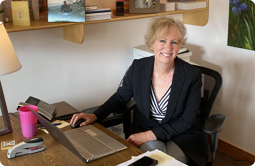 A smiling woman with short blond hair sits at a desk in a home office, using a laptop. Papers, a phone, a pink cup, and office supplies are on the desk. Shelves with photos and books are in the background.