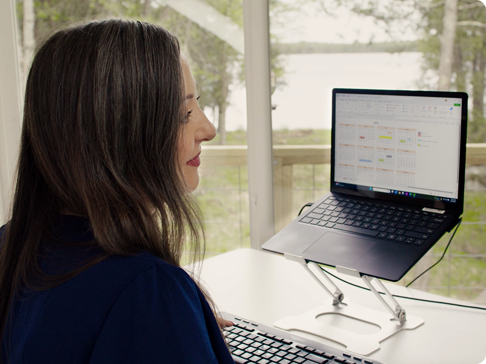 A remote worker with long dark hair works at a desk on a laptop placed on a stand. She uses a keyboard, focused on a spreadsheet, while large windows behind her reveal a view of trees and a lake.