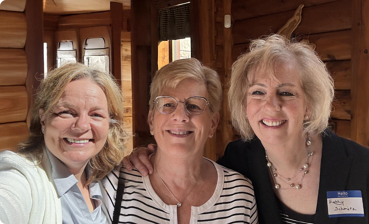 Three smiling women pose together indoors in a warmly lit, wooden room. The woman on the right wears a name tag reading Schwartz. All three appear happy and are close to each other, suggesting friendship or camaraderie.
