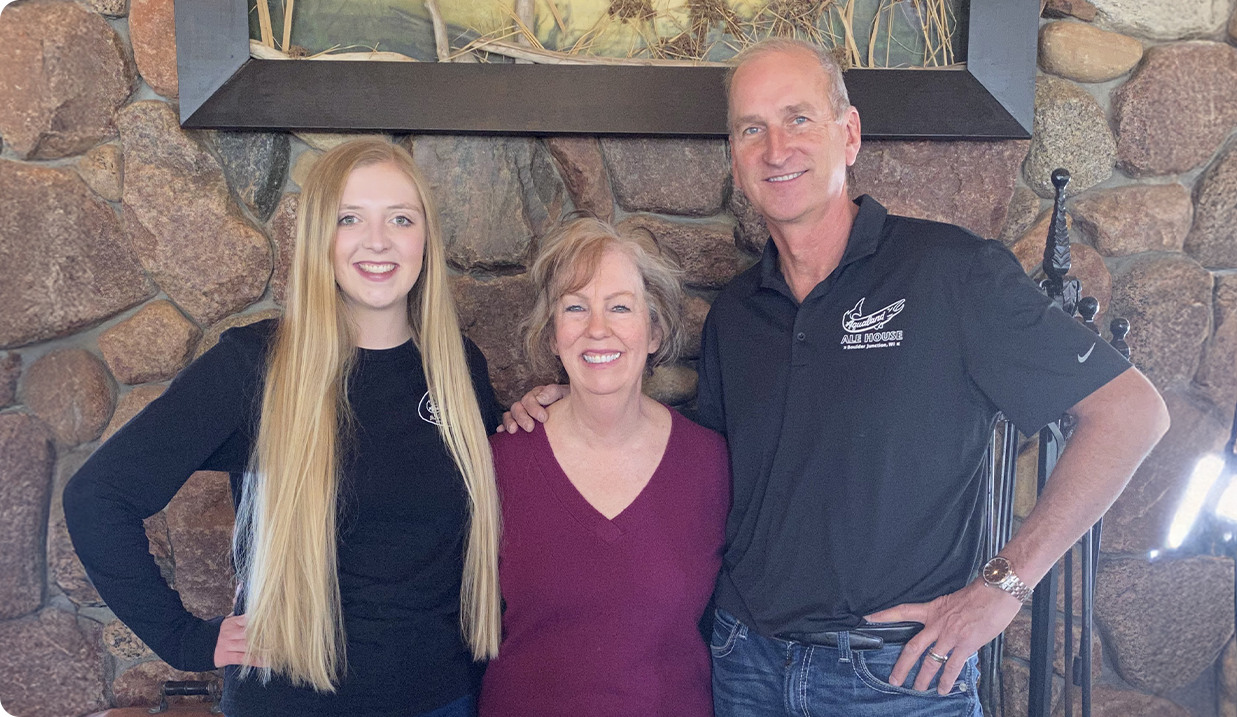 Three people stand smiling in front of a stone wall: a young woman with long blonde hair, a middle-aged woman in a maroon sweater, and a man in a black shirt with his arm around the middle woman.