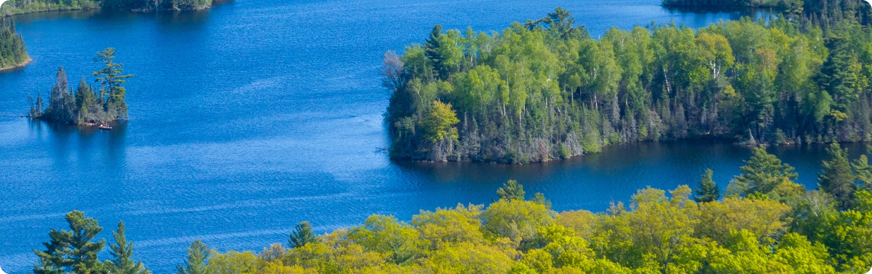 Aerial view of a blue lake with lush green, forested islands and shoreline under a bright sky, showcasing dense trees and calm, clear water.