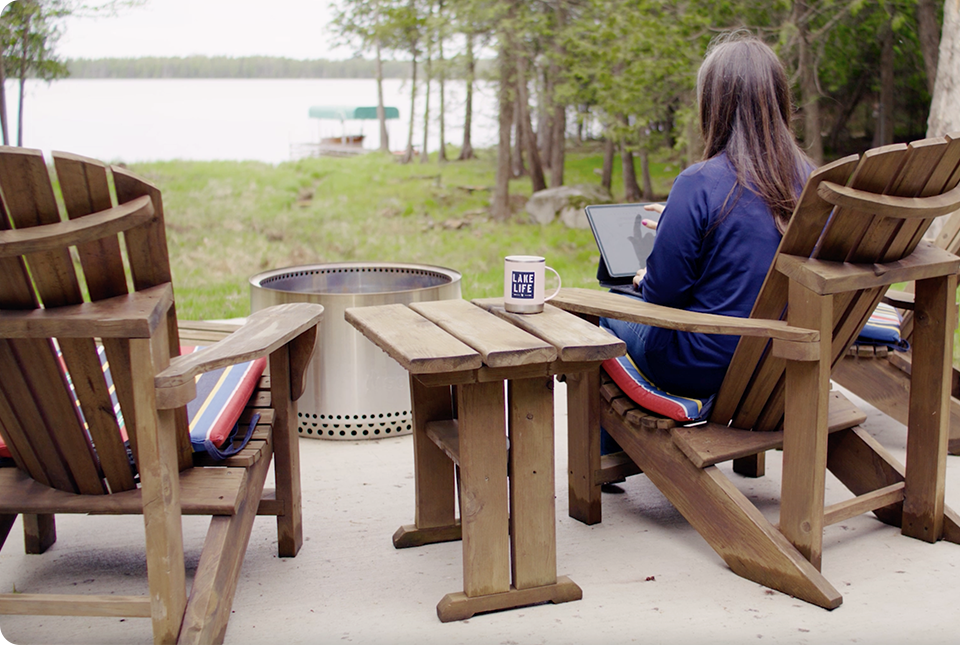 A person with long hair sits on a wooden chair outdoors, working on a laptop beside a small table and a fire pit, with a lake and trees visible in the background. A mug on the table reads LAKE LIFE.