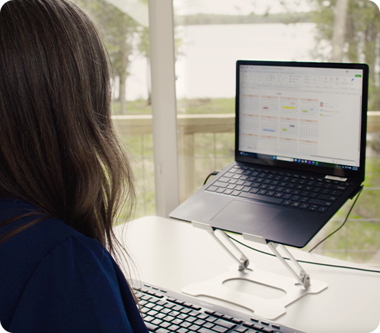 A remote worker with long brown hair sits at a desk, working on a laptop elevated on a stand. A spreadsheet is open on the screen, and there is a scenic outdoor view through large windows in the background.