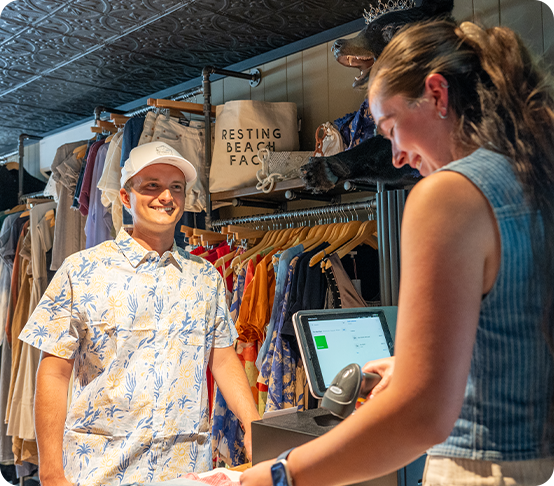 A smiling man in a floral shirt and white cap stands at a checkout counter while a cashier scans items in a boutique clothing store, reflecting the buzz of recent business expansion. Clothes and bags hang in the background.