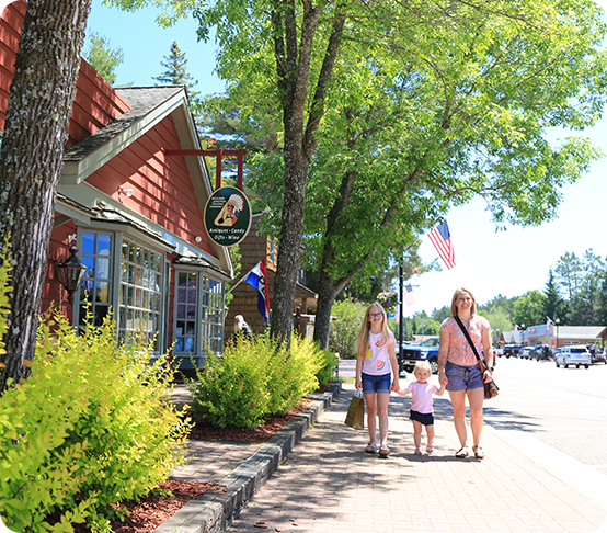 A woman and two children walk on a sunny sidewalk lined with trees and shops, reflecting recent business expansion. An American flag is displayed, several cars are parked along the street, and the sky is clear and blue.