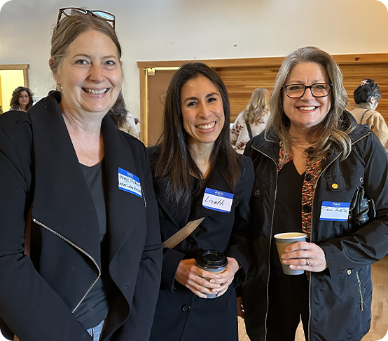 Three women wearing name tags smile at the camera while standing indoors. One, a remote worker, holds a coffee cup while another has a mug. Other people are visible in the background.