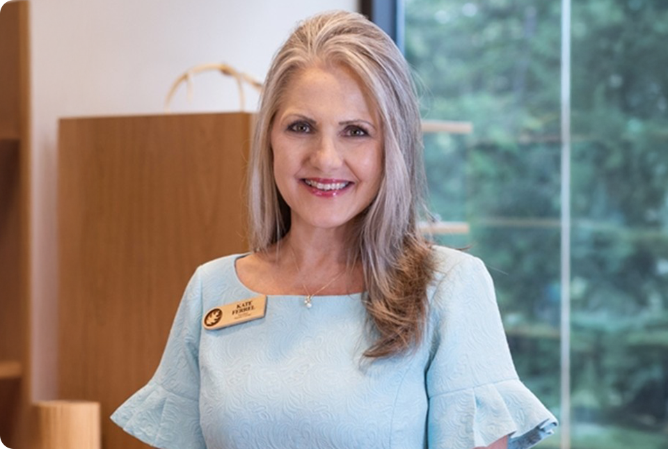 A smiling woman with long, blonde hair wearing a light blue dress and a name tag stands indoors, ready for a business expansion event, with a window and trees in the background.