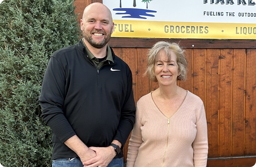 Two smiling entrepreneurs stand side by side outdoors in front of a wooden wall and a market sign. The man wears a black pullover; the woman wears a light pink sweater. A green bush is to the left.