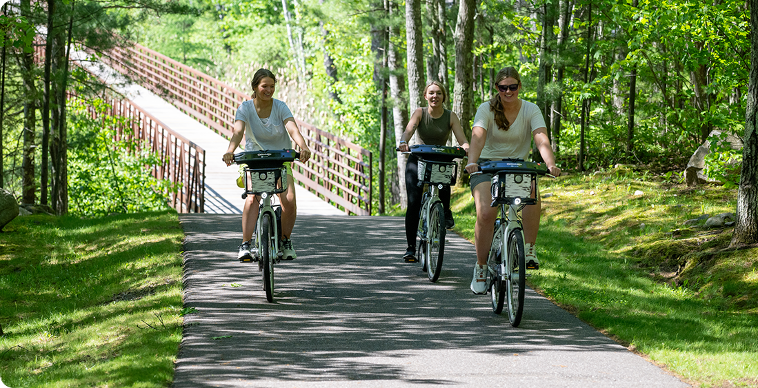 Three people ride bicycles along a paved path through a lush, green forest on a sunny day, smiling and enjoying the outdoors like a team celebrating business expansion, with a wooden bridge visible in the background.