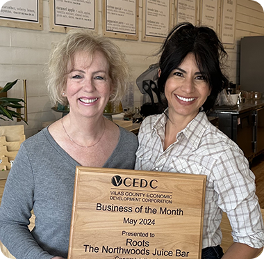 Two entrepreneurs smile while holding a plaque that reads Business of the Month, May 2024, presented to Roots The Northwoods Juice Bar, inside their juice bar with menus displayed on the wall behind them.