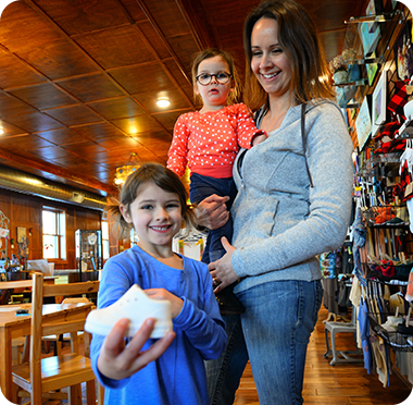 A smiling woman holds a young girl while another girl stands in front, smiling and holding a small white shoe in a cozy, well-lit startup store with wooden floors and shelves.