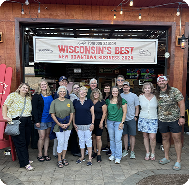 A group of fifteen smiling entrepreneurs pose together outside a building with a sign that reads, “Wisconsin’s Best New Downtown Business 2024” above the entrance. String lights hang overhead and a red Adirondack chair is on the left.