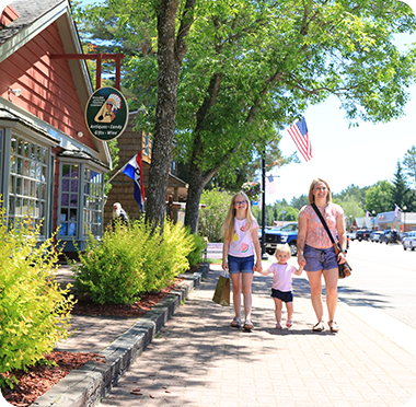 A woman and two children walk on a sunny, tree-lined sidewalk next to shops with red exteriors, where local entrepreneurs display their goods. An American flag hangs nearby, and cars are parked along the street in the background.