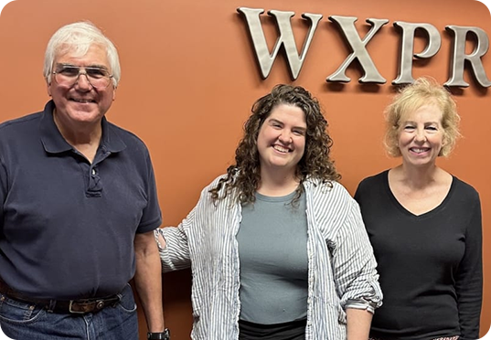 Three people stand smiling in front of an orange wall with large silver letters spelling WXPR. The two women are on the right, and the man is on the left.