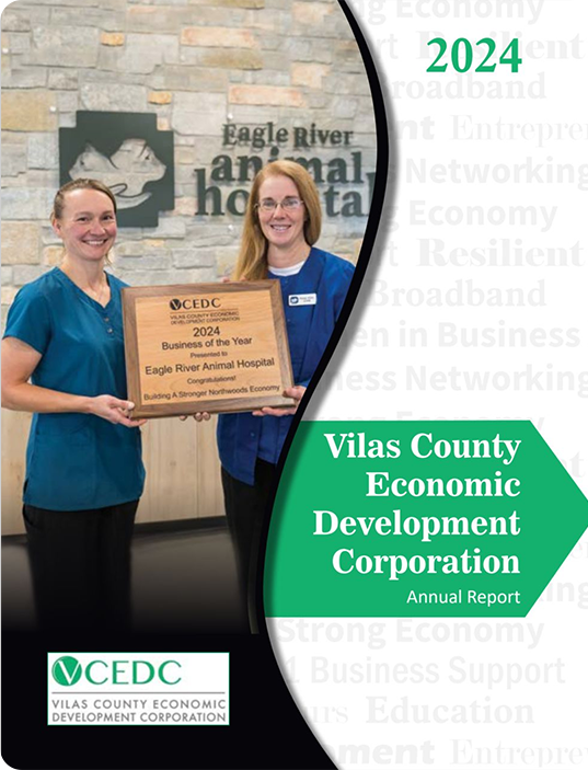 Two women stand indoors holding a plaque that reads 2024 Business of the Year, Eagle River Animal Hospital. The background features a sign for Eagle River Animal Hospital and the Vilas County Economic Development Corporation’s Annual Reports cover.