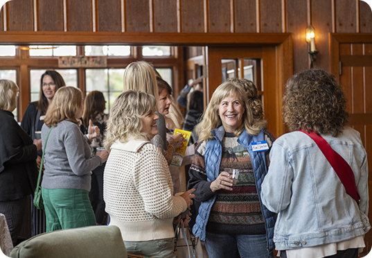 A group of women are standing and chatting at an events program in a cozy, wood-paneled room. Some are holding drinks. One woman in the foreground, smiling, wears a blue vest and name tag. Warm light fills the space.
