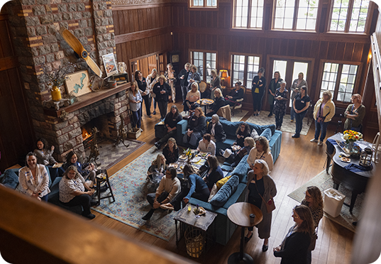 A large group of people gathers in a cozy, wood-paneled living room with a stone fireplace, sofas, and snacks, enjoying conversation during an engaging program or events as natural light streams in from tall windows.