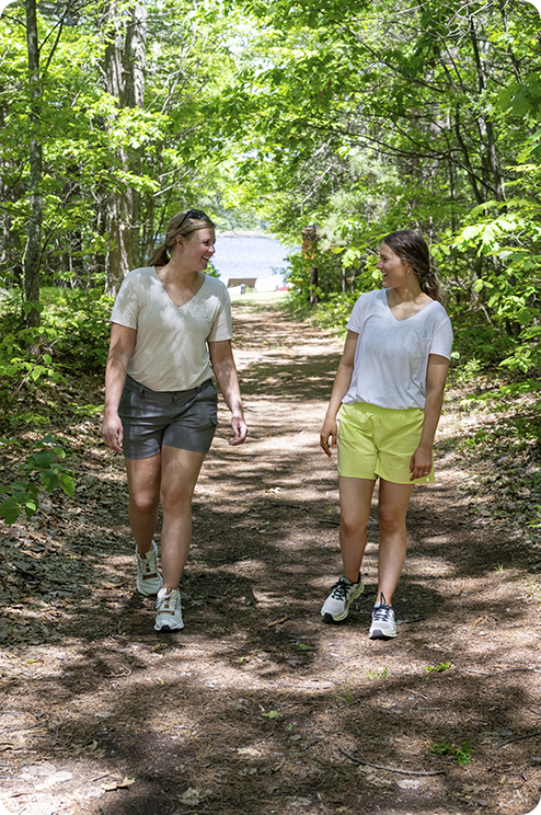 Two women walk and talk on a sunlit forest trail surrounded by green trees. Both wear shorts, T-shirts, and sneakers, enjoying a summer day outdoors. A lake is visible in the background through the trees.