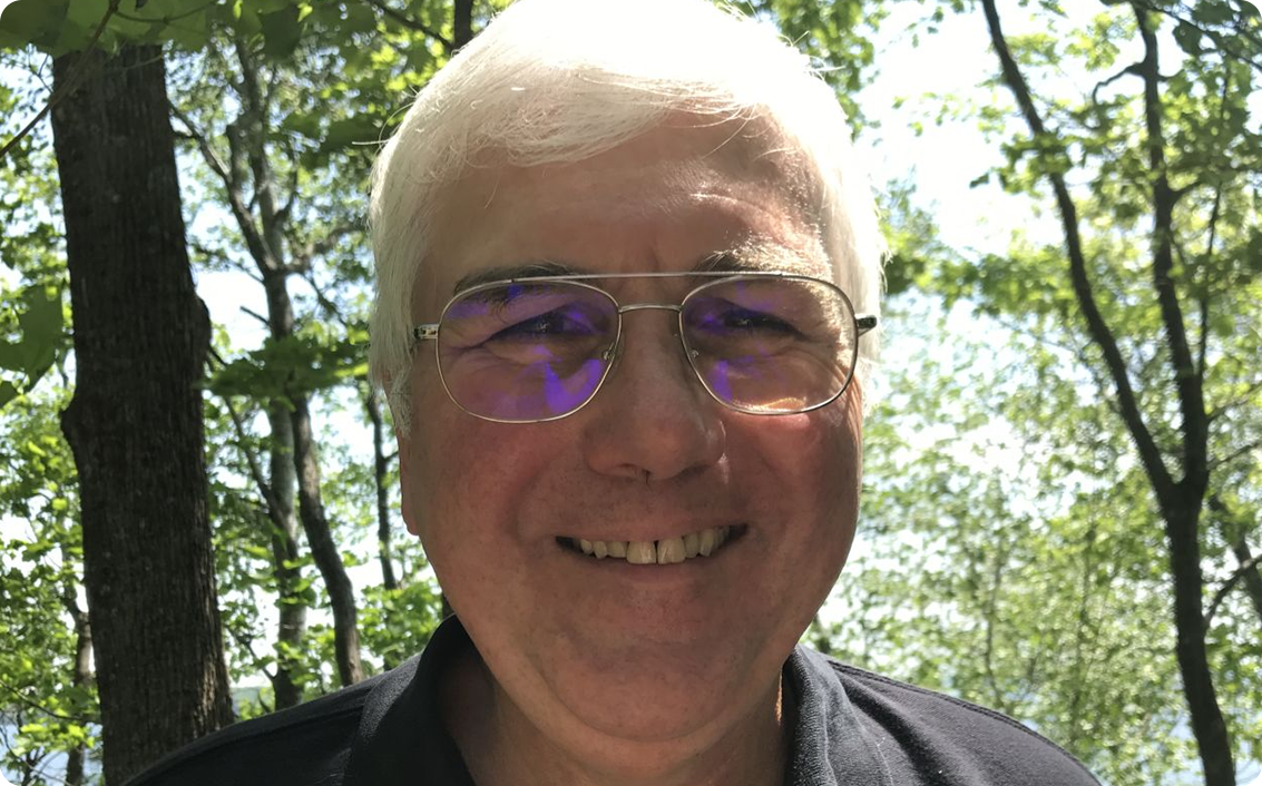 A smiling older man with white hair and glasses stands outdoors among green trees on a sunny day, reflecting the optimism that comes with wise investment.