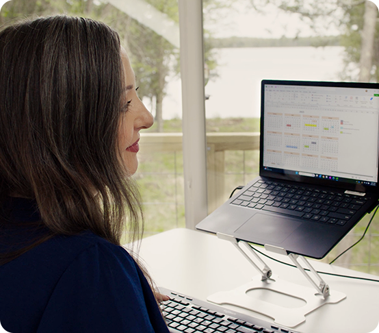A woman with long dark hair works at a desk using a keyboard and a laptop on a stand. She is facing the screen, which displays charts. Large windows behind her show a scenic outdoor view with trees and water.