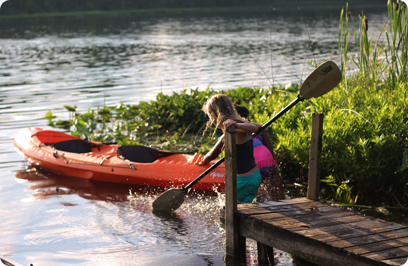 Two children stand on a wooden dock beside a lake, using paddles to splash water near an orange kayak partially in the water, surrounded by green plants on a calm, sunny day.