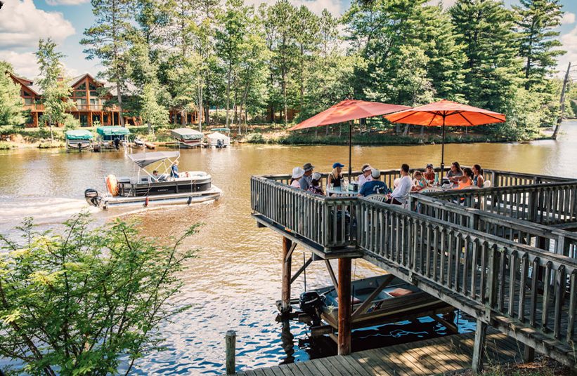 People sit under orange umbrellas on a wooden deck overlooking a river, while a pontoon boat passes by. The river is surrounded by trees and lakeside houses, creating a peaceful, summery atmosphere.