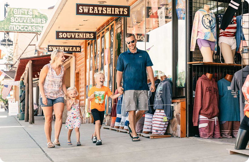 A smiling family of four, two adults and two young children, walk hand in hand past a souvenir and clothing shop on a sunny day. Mannequins and signs for sweatshirts and gifts are visible in the shop window.