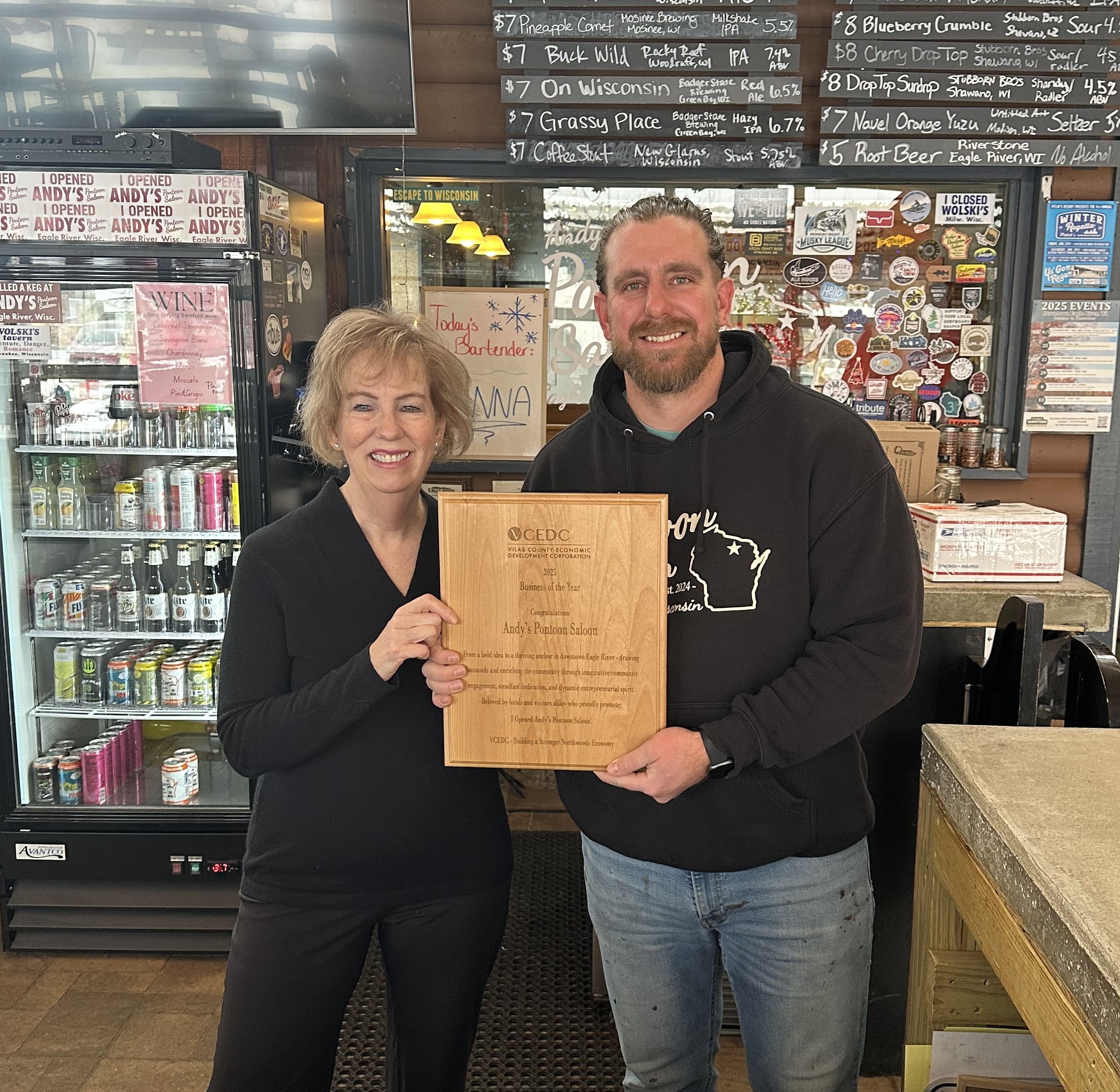 Two people stand indoors holding a wooden award plaque. They are smiling at the camera. Behind them are a beverage cooler, a chalkboard menu, and a wall decorated with various stickers.
