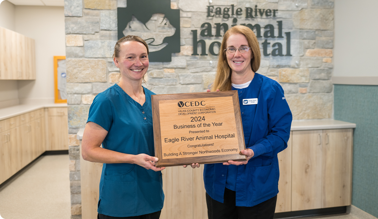 Two women in blue uniforms smile and hold a plaque that reads, “2024 Business of the Year presented to Eagle River Animal Hospital.” They stand in a modern veterinary clinic with a stone wall and hospital sign behind them.