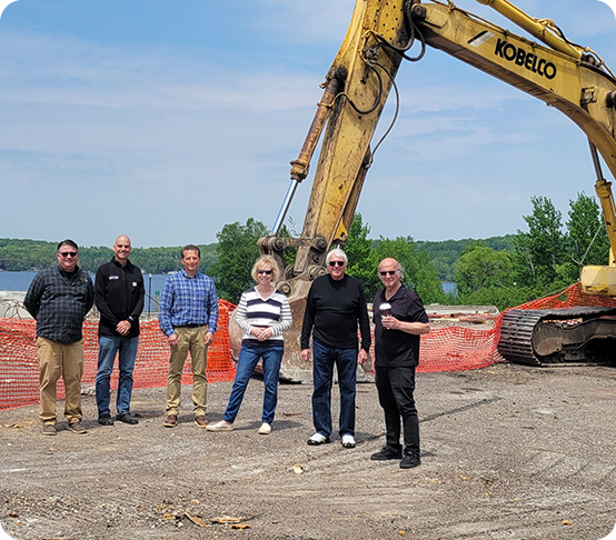 Six entrepreneurs stand in front of an excavator at a construction site near a lake, with orange safety fencing and green trees in the background under a blue sky.