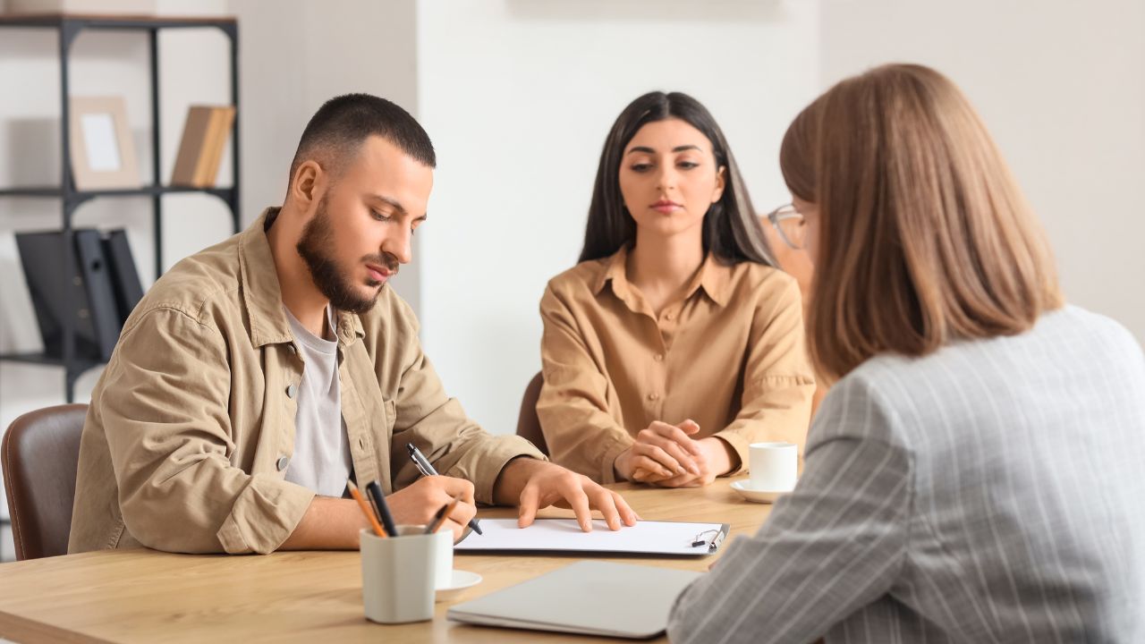 A man is signing a document at a desk while a woman, representing Women in Business, sits beside him; both face a Program Manager with a notepad during a meeting in a bright office setting.