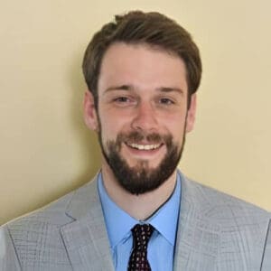 A man with brown hair and a beard smiles at the camera. He is wearing a light gray suit jacket, light blue shirt, and a patterned dark tie, ready to discuss business taxes while standing against a plain beige background.