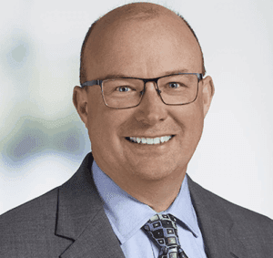 A smiling middle-aged man with glasses, wearing a gray suit, blue shirt, and patterned tie, poses in front of a blurred light background—projecting confidence ideal for topics like business loans or business grants.