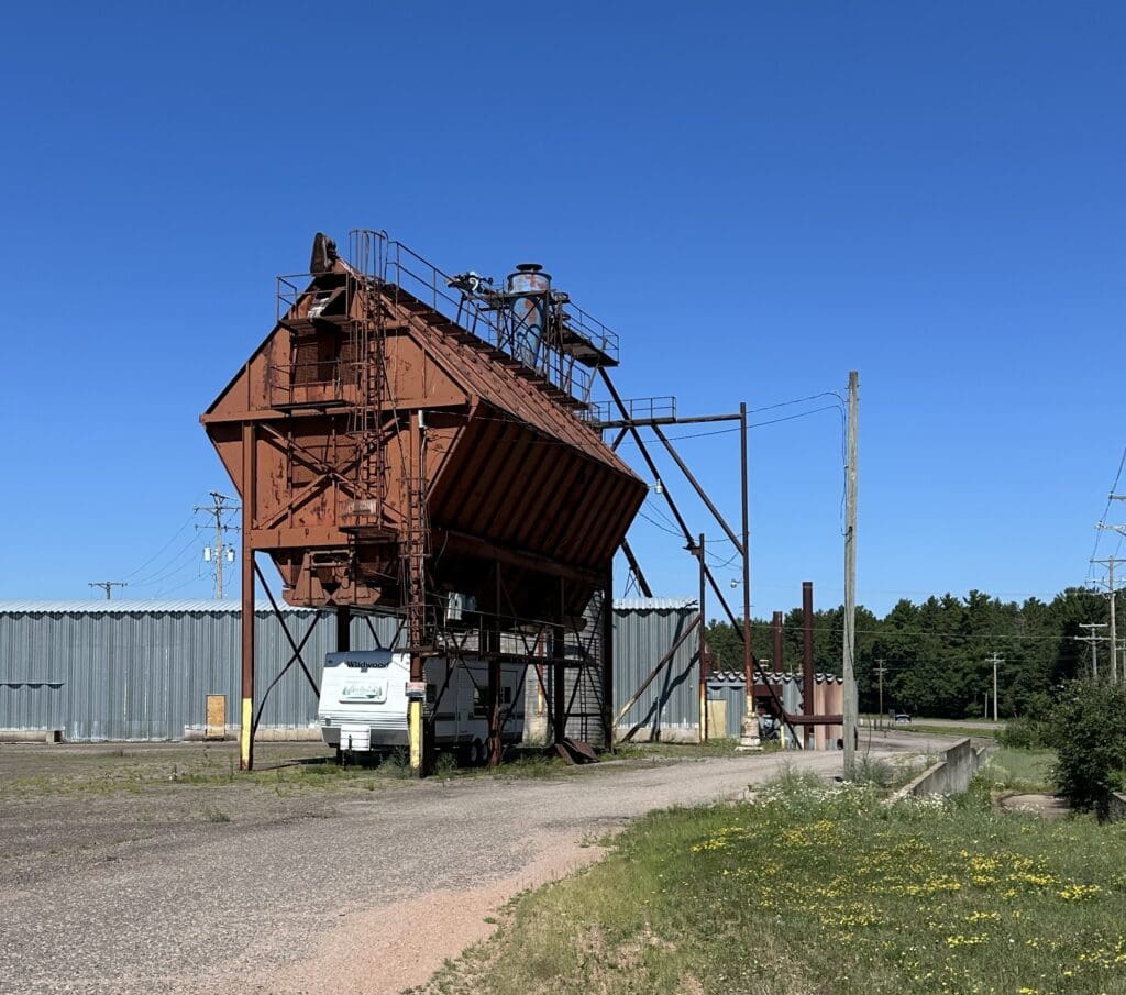 Large rust-colored industrial structure on stilts, with a white truck parked underneath. Set against the blue sky and grassy area typical of rural Wisconsin, utility poles dot the landscape. A metal warehouse and trees are visible in the background, reflecting the growth spurred by initiatives like the WEDC Thrive Rural Wisconsin Grant and VCEDC support.